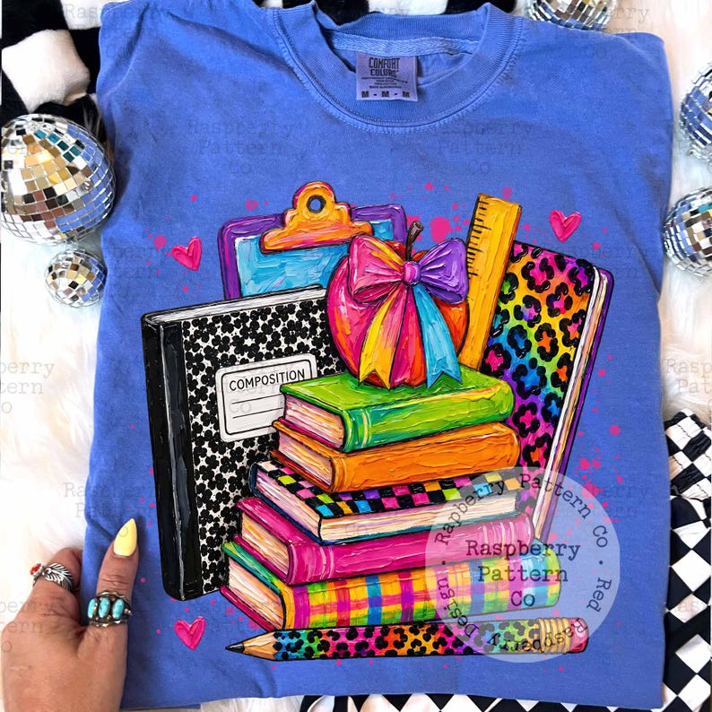 May include: A periwinkle blue t-shirt featuring a colorful graphic of a stack of books, a composition notebook, a clipboard, a ruler, and a pencil. The design includes a bright apple with a rainbow bow, leopard print accents, and small hearts.