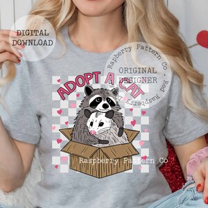 Op de afbeelding: Een grijs T-shirt met een afbeelding van een wasbeer en een opossum in een kartonnen doos. De tekst "Adopt a Cat" staat boven de dieren. De tekst "Raspberry Pattern Co." staat onder de dieren.