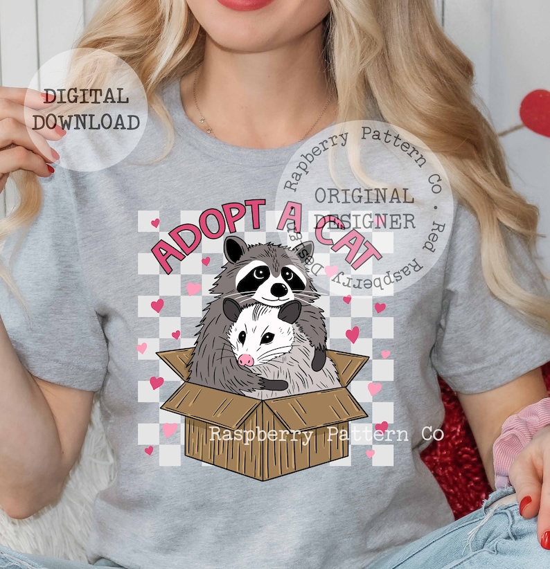 Op de afbeelding: Een grijs T-shirt met een afbeelding van een wasbeer en een opossum in een kartonnen doos. De tekst "Adopt a Cat" staat boven de dieren. De tekst "Raspberry Pattern Co." staat onder de dieren.
