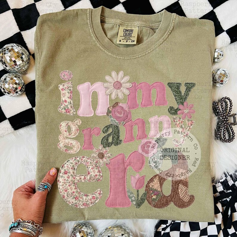 May include: Olive green Comfort Colors t-shirt with the phrase "in my granny era" appliqued in pink and floral fabrics. The design includes flowers and a tulip. The shirt has a small tag that says "S-P-CH".