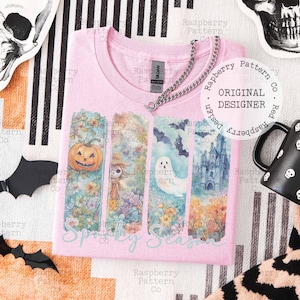 May include: Pink t-shirt with a "Spooky Season" graphic. The design features a pumpkin, scarecrow, ghost, bats, and a haunted castle. A silver chain necklace is draped across the shirt.