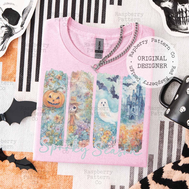 May include: Pink t-shirt with a "Spooky Season" graphic. The design features a pumpkin, scarecrow, ghost, bats, and a haunted castle. A silver chain necklace is draped across the shirt.