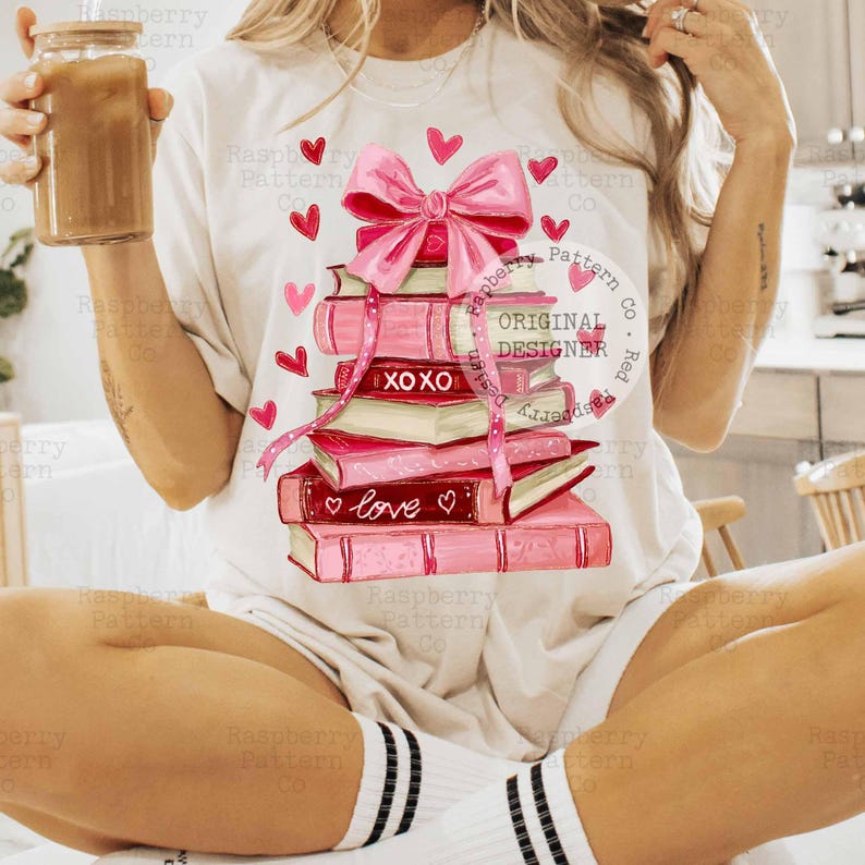 May include: White t-shirt featuring a stack of pink books with a large pink bow and hearts. The books have the words "XOXO" and "love" on them. The shirt is a casual style.