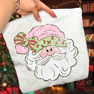 May include: A white pillow with a Santa Claus design. The Santa has a pink hat and a green hatband with red berries. A large bow with red stripes and green accents is on the hat. The design is outlined in gold.