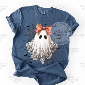 K&ouml;nnte beinhalten: Ein blaues T-Shirt mit einem skurrilen Geisterdesign. Der Geist hat eine pfirsichfarbene Schleife, schwarze Augen und einen Spitzenbesatz. Ein Spinnennetz und Flederm&auml;use umgeben den Geist. Das Shirt hat den Text "ORIGINAL DESIGNER".