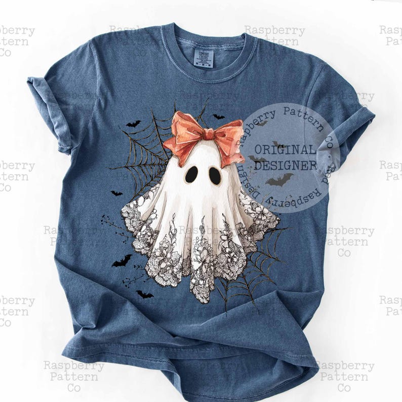 K&ouml;nnte beinhalten: Ein blaues T-Shirt mit einem skurrilen Geisterdesign. Der Geist hat eine pfirsichfarbene Schleife, schwarze Augen und einen Spitzenbesatz. Ein Spinnennetz und Flederm&auml;use umgeben den Geist. Das Shirt hat den Text "ORIGINAL DESIGNER".