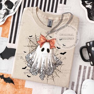 K&ouml;nnte beinhalten: Beiges T-Shirt mit Geisterdesign, korallenfarbener Schleife, schwarzer Spitze und Spinnweben. Das Shirt hat einen Rundhalsausschnitt und kurze &Auml;rmel. Das Design beinhaltet Flederm&auml;use und den Text "Original Designer".