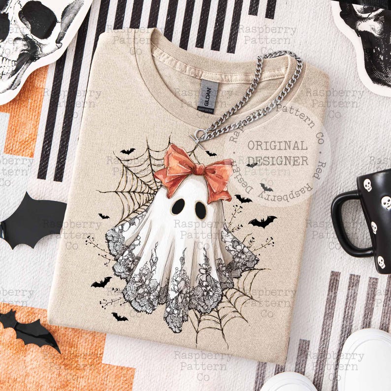 K&ouml;nnte beinhalten: Beiges T-Shirt mit Geisterdesign, korallenfarbener Schleife, schwarzer Spitze und Spinnweben. Das Shirt hat einen Rundhalsausschnitt und kurze &Auml;rmel. Das Design beinhaltet Flederm&auml;use und den Text "Original Designer".
