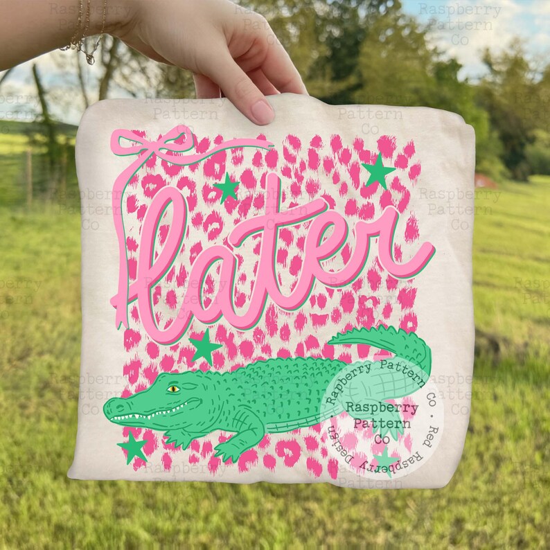 May include: A beige tote bag featuring a pink leopard print background with the word "Later" in pink script. A green alligator graphic is at the bottom, with green star accents and a pink bow at the top. The bag is held by a person.