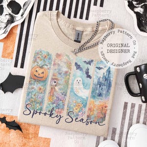 May include: Beige t-shirt with a colorful "Spooky Season" graphic. The design features a pumpkin, scarecrow, ghost, and castle. The shirt has a crew neck and short sleeves. A silver chain necklace is draped on the shirt.