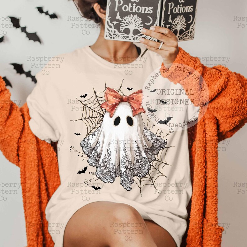 K&ouml;nnte beinhalten: Cremefarbenes T-Shirt mit einem Aquarell-Geisterdesign, einer roten Schleife, schwarzen Augen und Spitzendetails. Der Geist ist von einem Spinnennetz und Flederm&auml;usen umgeben. Der Text "Raspberry Pattern Co" ist sichtbar.
