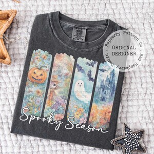 May include: Dark gray t-shirt with a "Spooky Season" graphic. The design features watercolor illustrations of a jack-o'-lantern, scarecrow, ghost, and haunted castle. The shirt has a crew neck and short sleeves.