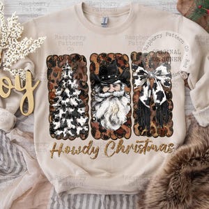 May include: Beige sweatshirt with a festive design. Features three rectangular panels with a Christmas tree, Santa Claus, and a bow, all with a cowhide pattern. The text "Howdy Christmas" is below the panels.