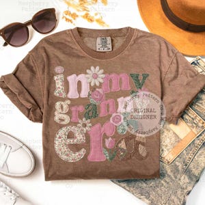 May include: A brown t-shirt with the phrase "in my grandma era" in floral and pink applique letters. The shirt is styled with sunglasses, a hat, and jeans.