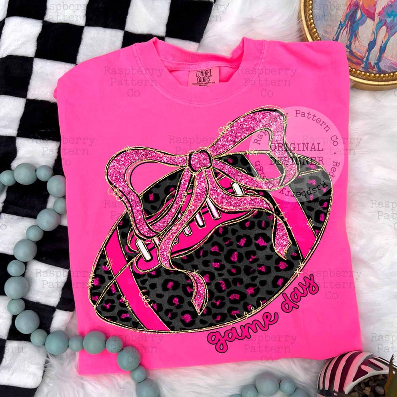 May include: A bright pink t-shirt featuring a football design with a pink glitter bow and leopard print accents. The football has pink stripes and the words "game day" in a script font. The shirt is a Comfort Colors brand.