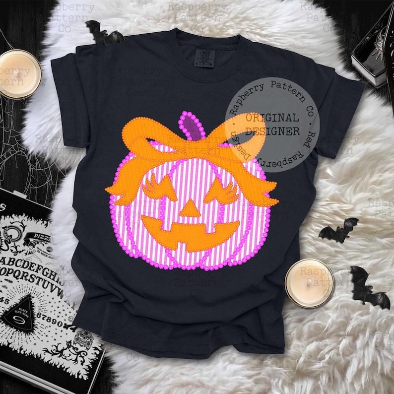 以下が含まれることがあります： ハロウィーンをテーマにしたデザインの黒いTシャツ。デザインには、オレンジ色のリボンと顔が付いた、ピンクと白のストライプのかぼちゃが含まれています。シャツには「Raspberry Pattern Co」と「Original Designer」の文字が入っています。