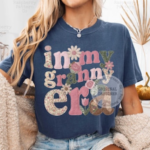 May include: Navy blue t-shirt with the phrase "in my granny era" in a floral and patchwork design. The letters are in pink, green, and floral patterns, with flower accents. A casual, vintage-inspired top.