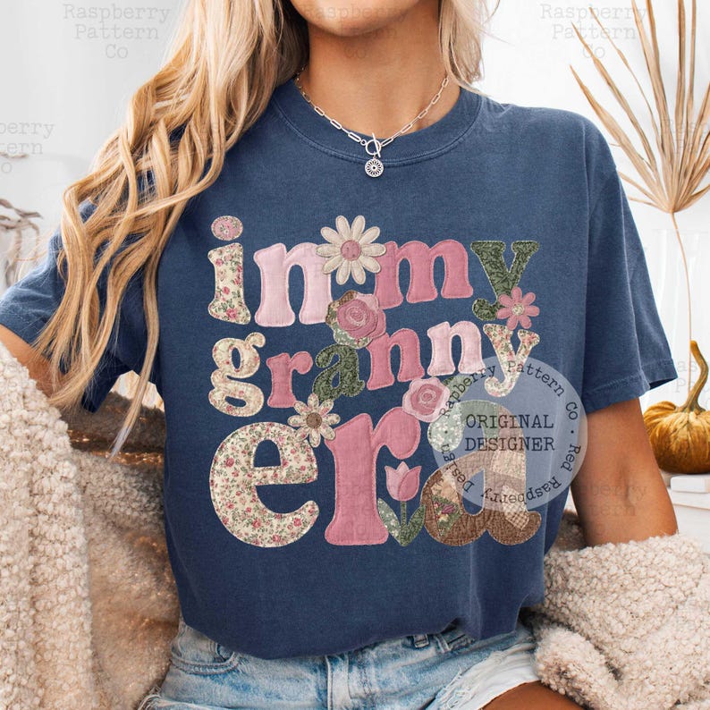 May include: Navy blue t-shirt with the phrase "in my granny era" in a floral and patchwork design. The letters are in pink, green, and floral patterns, with flower accents. A casual, vintage-inspired top.