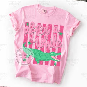 May include: Pink t-shirt with the text "After a While" in a script font, over pink and white stripes. A green alligator graphic is below the text, with green stars. The shirt is a casual style.
