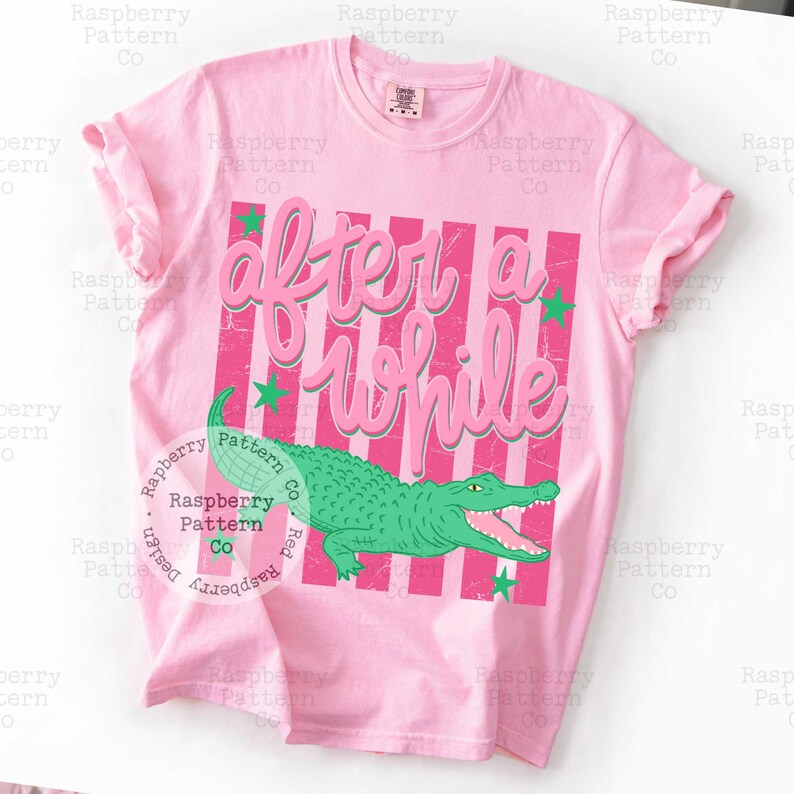 May include: Pink t-shirt with the text "After a While" in a script font, over pink and white stripes. A green alligator graphic is below the text, with green stars. The shirt is a casual style.