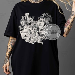 May include: Black t-shirt featuring a vintage-style graphic with an angel holding a heart, surrounded by floral elements, the words "BE MINE", and "XOXO". The design includes the text "kisses" and "ORIGINAL DESIGNER".