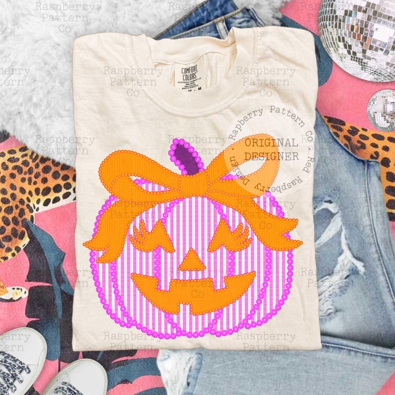 以下が含まれることがあります： ハロウィーンをテーマにしたデザインのクリーム色のTシャツ。デザインには、オレンジ色のリボンと顔が付いた、ストライプの紫と白のカボチャが含まれています。「ORIGINAL DESIGNER」という文字がTシャツに表示されています。