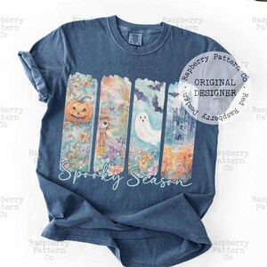 May include: A blue t-shirt featuring a colorful, vertical design with a pumpkin, scarecrow, ghost, and haunted house. The words "Spooky Season" are written in a script font below the design. A circular logo reads "Original Designer".
