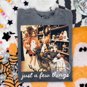 May include: A charcoal gray crewneck t-shirt featuring a graphic print of a doll in an orange sweater pushing a shopping cart filled with Halloween decor. The text "WITCH BETTER HAVE MY CANDY" and "just a few things" is printed on the shirt.
