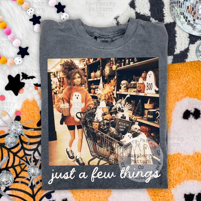 May include: A charcoal gray crewneck t-shirt featuring a graphic print of a doll in an orange sweater pushing a shopping cart filled with Halloween decor. The text "WITCH BETTER HAVE MY CANDY" and "just a few things" is printed on the shirt.