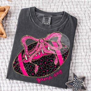 May include: Dark gray t-shirt featuring a pink football design with a leopard print pattern and a glittery pink bow. The words "game day" are written in pink script. The shirt has a crew neck and short sleeves.