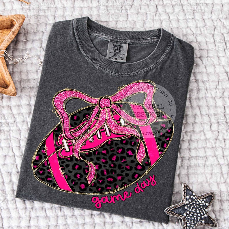 May include: Dark gray t-shirt featuring a pink football design with a leopard print pattern and a glittery pink bow. The words "game day" are written in pink script. The shirt has a crew neck and short sleeves.