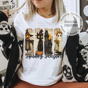 May include: White t-shirt featuring a Halloween-themed design. The graphic includes a scarecrow, black cat, haunted house, and ghost, with the words "Spooky Season" below. The design is in a vintage style.