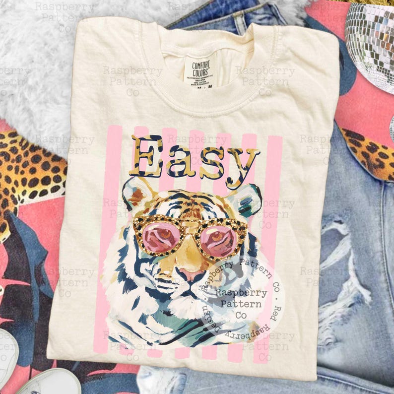 May include: Cream-colored t-shirt with a tiger graphic wearing pink sunglasses. The word "Easy" is printed above the tiger in a leopard print font, set against pink vertical stripes. The shirt is part of a clothing collection.