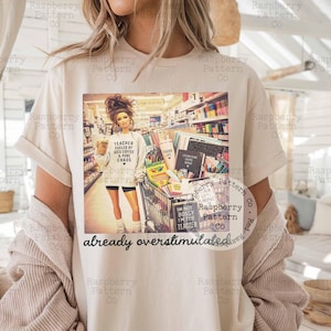 May include: A beige t-shirt featuring a graphic of a person in a store aisle with a shopping cart full of school supplies. The graphic includes the text "TEACHER FUELED BY ICED COFFEE & PURE CHAOS" and "already overstimulated" printed below the image.