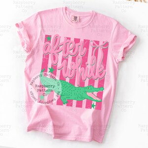 May include: Pink t-shirt with the text "After While" in a pink script font over a pink and white striped background. A green alligator graphic is below the text, with green star accents.