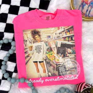 May include: A neon pink crewneck t-shirt featuring a graphic print of a person in a classroom setting with a shopping cart full of school supplies. The person wears a shirt that reads Teacher Fueled By Iced Coffee & Pure Chaos.