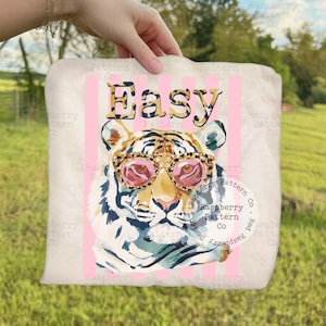 May include: A white pillow with a tiger wearing pink sunglasses and the word "Easy" in a leopard print font. Pink vertical stripes are behind the tiger. The pillow is being held by a person.