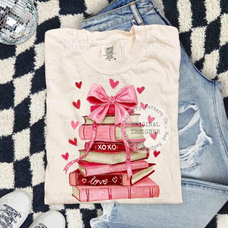 May include: Cream-colored t-shirt featuring a stack of pink books with a large pink bow and hearts. The books have the words "XOXO" and "love" on them. The shirt is on a black and white checkered surface with jeans and white sneakers.