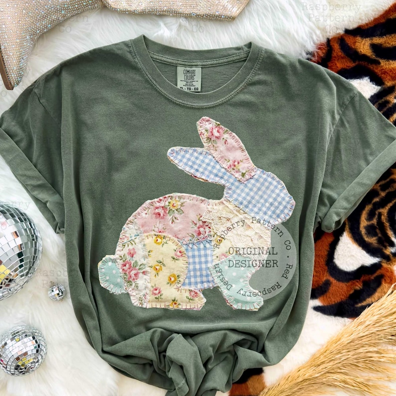 May include: Olive green t-shirt featuring a patchwork bunny design. The bunny is composed of floral, gingham, and polka dot fabrics. The text "Raspberry Pattern Co. Original Designer" is printed on the shirt.