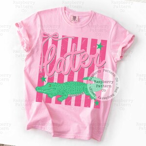May include: A light pink t-shirt with a graphic design. The design features the word "Later" in pink, a green alligator, pink and white stripes, and green star accents. The shirt is a crew neck.