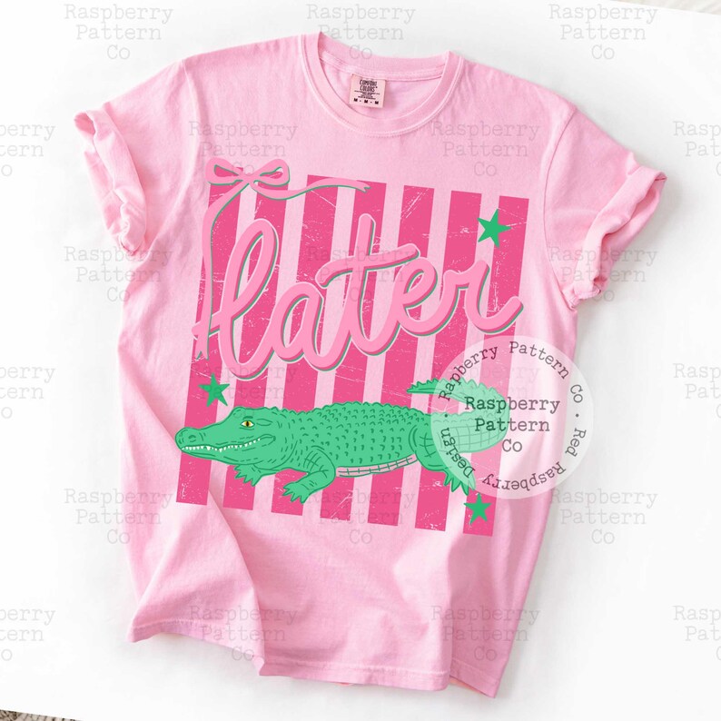 May include: A light pink t-shirt with a graphic design. The design features the word "Later" in pink, a green alligator, pink and white stripes, and green star accents. The shirt is a crew neck.