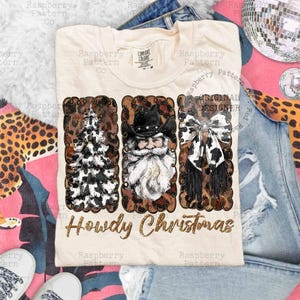 May include: Cream-colored t-shirt with a festive design. The design features a Christmas tree, Santa Claus in a cowboy hat, and a cow-print bow, all framed with a leopard print border. The text "Howdy Christmas" is written in gold glitter.