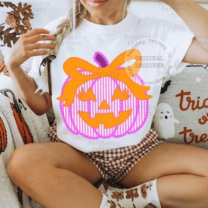 以下が含まれることがあります： ハロウィーンをテーマにしたデザインの白いTシャツ。デザインには、ピンクと白のストライプのボディ、紫の茎、オレンジ色のリボンが付いた、笑顔のオレンジ色のカボチャが含まれています。「ORIGINAL DESIGNER」という文字が見えます。