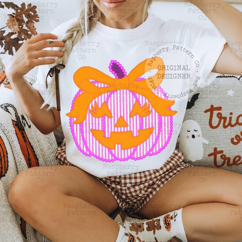 以下が含まれることがあります： ハロウィーンをテーマにしたデザインの白いTシャツ。デザインには、ピンクと白のストライプのボディ、紫の茎、オレンジ色のリボンが付いた、笑顔のオレンジ色のカボチャが含まれています。「ORIGINAL DESIGNER」という文字が見えます。