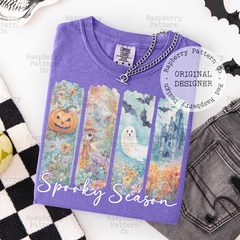 May include: A purple V-neck t-shirt with a "Spooky Season" graphic. The graphic features a pumpkin, scarecrow, ghost, and castle. A silver chain necklace is draped on the shirt. The shirt is made of Comfort Colors material.