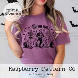 Puede incluir: Una camiseta morada con un dise&ntilde;o gr&aacute;fico negro de un esqueleto, un vampiro y un monstruo de Frankenstein bailando. El texto en la camiseta dice "It was a Graveyard Smash".