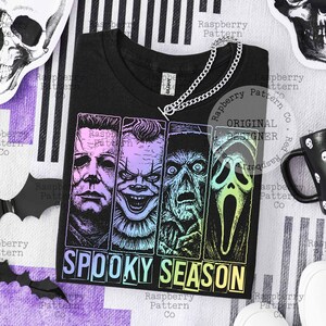 Peut inclure: T-shirt noir avec un motif de quatre personnages de films d'horreur et l'inscription "SPOOKY SEASON" en d&eacute;grad&eacute;. Un collier cha&icirc;ne argent&eacute; est pos&eacute; sur le t-shirt. Le t-shirt est sur un fond ray&eacute; avec des d&eacute;corations d'Halloween.