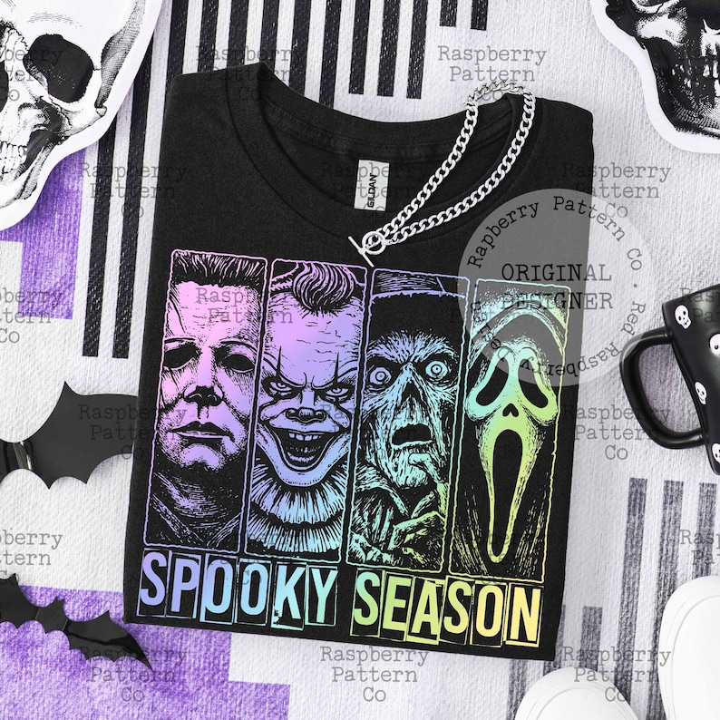 Peut inclure: T-shirt noir avec un motif de quatre personnages de films d'horreur et l'inscription "SPOOKY SEASON" en d&eacute;grad&eacute;. Un collier cha&icirc;ne argent&eacute; est pos&eacute; sur le t-shirt. Le t-shirt est sur un fond ray&eacute; avec des d&eacute;corations d'Halloween.