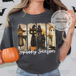 May include: Dark gray t-shirt featuring a Halloween-themed design. The design includes a scarecrow, black cat, haunted house, and ghost, with the words "Spooky Season" in white script. The shirt has a vintage look.