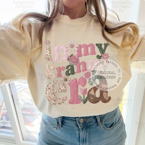 May include: Cream-colored sweatshirt with the phrase "in my grandma era" in a floral and pink design. The letters are decorated with flowers and patterns. The sweatshirt is paired with blue jeans.